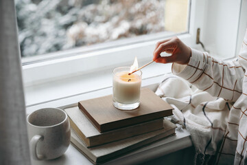 Kids hand in checkered pyjamas with burning match lighting candle on windowsill. Autumn still life with cup of coffee, oldbooks. Autumn home decor. Blurred garden background. Cozy fall, winter mood.
