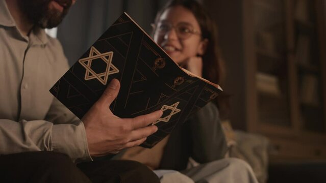 Close-up selective focus shot of hands of anonymous Jewish man holding holy Torah book, with star of David on cover, while sitting on sofa at home and discussing scriptures with teenage daughter