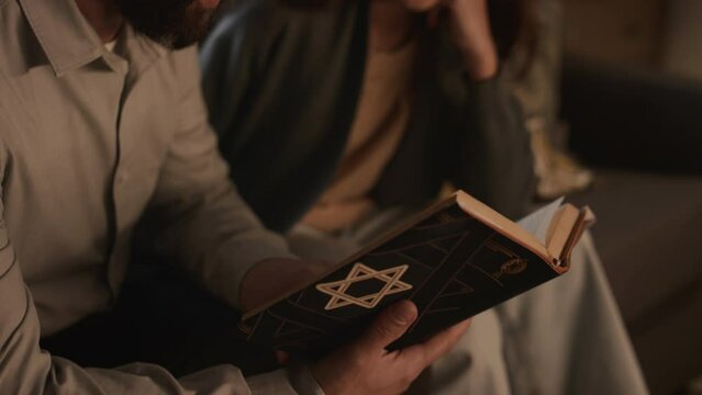 Close-up tilting down shot of Jewish father with beard, wearing yarmulke skullcap, sitting on couch with teenage daughter, holding Torah with star of David, reading and discussing holy texts together