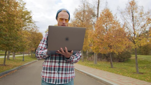 Male tourist with black backpack with red beard in yellow sun glasses typing on silver laptop, standing on asphalt bicycle path with yellow trees on sides. High quality 4k footage
