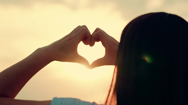 Young woman draws heart hands against sky. Silhouette of heart hands in sun close-up. Girl make sign love hands sunset. Hands in shape heart. Sunlight between fingers. Sign, heart symbol love, feeling