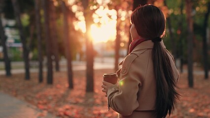 Young woman walks in autumn city park with mug of coffee. Beautiful fashionable girl drinks coffee...