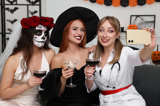 Group of women in scary costumes with cocktails taking selfie at Halloween party indoors
