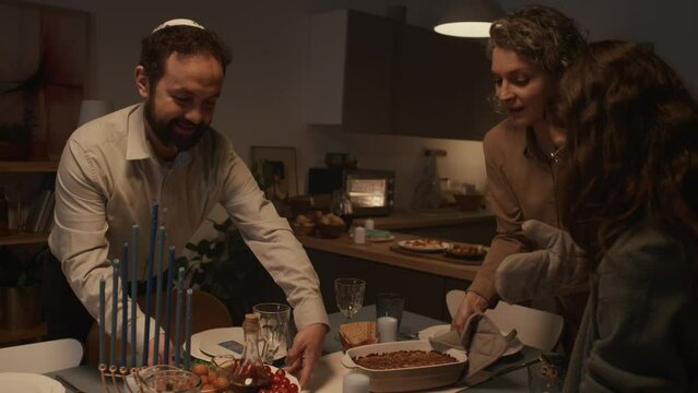 Medium shot of Jewish mother, father, daughter preparing dinner table for family feast on Hanukkah - laying down plates with food and hot kugel, looking around, chatting with pleasure and excitement