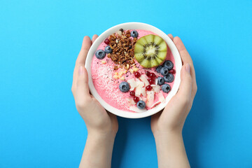 Woman holding tasty smoothie bowl with fresh kiwi fruit, berries and granola at light blue table,...