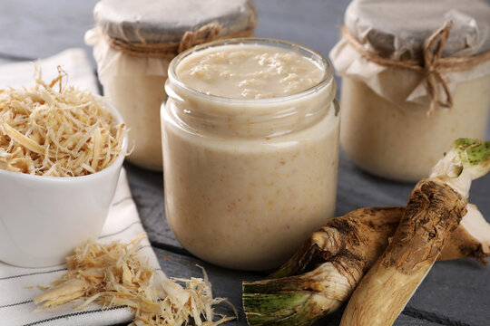 Spicy horseradish sauce in jars and roots on grey wooden table, closeup