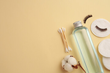 Bottle of makeup remover, cotton flower, pads, swabs and false eyelashes on yellow background, flat lay. Space for text