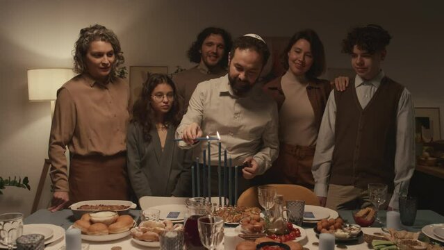 Medium shot of Jewish family of six standing around dinner table at Hanukkah celebration, man in kippah holding burning shammash and lighting up candles on hanukkiah, while everybody is watching