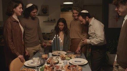Full shot of teenage Jewish girl in glasses standing at dinner table with family members, lighting up candles on hanukkiah with shammash, while celebrating on final day of Hanukkah religious festival
