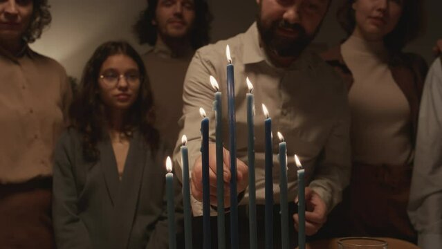 Medium shot of middle-aged Jewish man in kippah skullcap lighting up eight Hanukkah candles on menorah using shammash, then putting it in middle, singing and dancing with joy