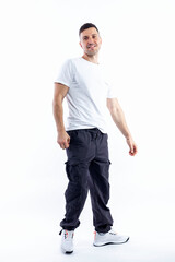 A handsome young man stands tall. A smiling brunette guy in a white T-shirt, dark trousers and sneakers. Positive and optimistic. Isolated on a white background. Vertical. Space for text.