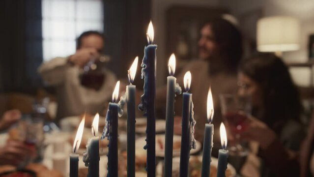 Close-up selective focus shot of nine burning candles on menorah on final day of Hanukkah holliday, and Jewish family of six having dinner together, smiling and talking in blurred background