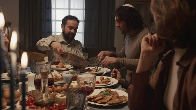 Medium shot of cheerful Jewish family eating dinner together on final day of Hanukkah, smiling, chatting, father in kippah holding tray, son taking out piece of kugel and laughing