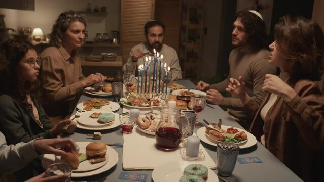 Medium shot of Jewish family of six sitting around festive table, eating dinner to celebrate Hanukkah, young woman telling funny story with energetic gestures and everybody listening