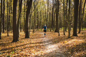 Fototapeta premium Young man jogging outdoors in a sunny autumn forest. Fitness male exercising in the park, wearing blue and black sportswear. People and sport concept healthy lifestyle, self-love and wellness.