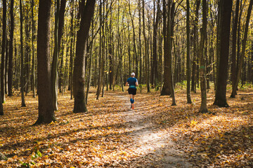 Young man jogging outdoors in a sunny autumn forest. Fitness male exercising in the park, wearing blue and black sportswear. People and sport concept healthy lifestyle, self-love and wellness.