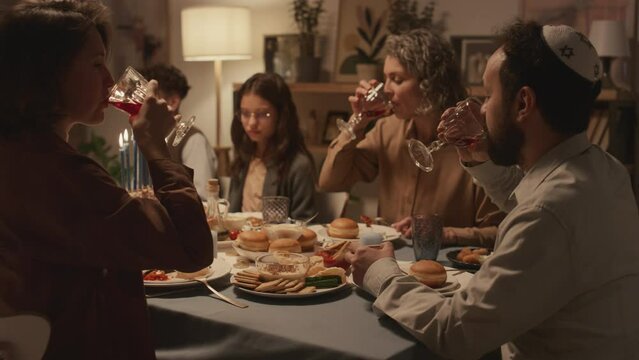 Medium shot of large Jewish family enjoying dinner together to celebrate Hanukkah, eating traditional foods and snacks, talking, smiling, raising glasses and toasting each other