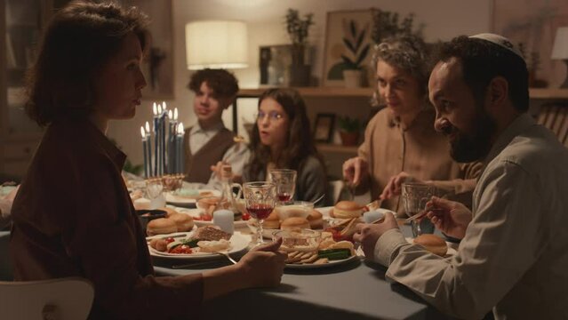 Medium shot of Jewish family sitting around festive table and enjoying traditional foods at Hanukkah dinner, dad in kippah talking to female relative or friend, then both laughing
