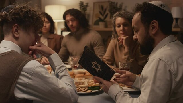 Medium shot of Jewish man in kippah sitting at Hanukkah dinner with family and friends, reciting solemn passages from holy Torah book with star of David on cover, and people sitting and listening