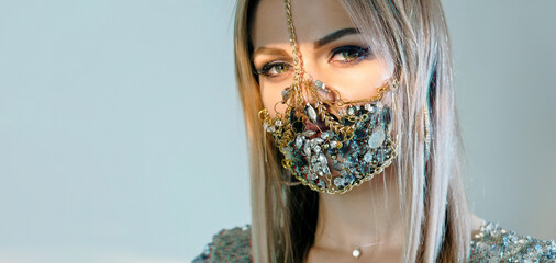 Pandemic fashion. Quarantine DIY accessories. Portrait of sensual woman in glamour handmade chain gemstone face mask posing isolated on blue copy space background.