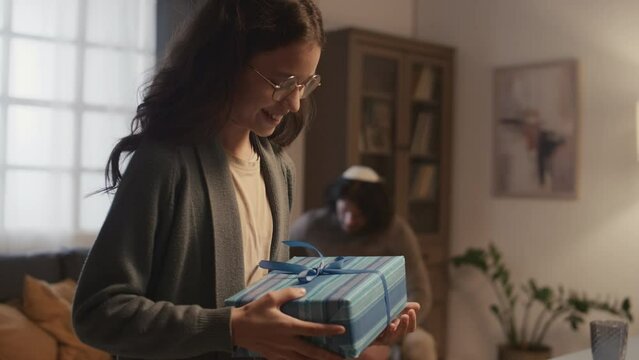 Medium close-up shot of teenage Jewish girl in glasses receiving present in blue box from her aunt, while celebrating Hanukkah with family at home, and man in kippah sitting on couch in background