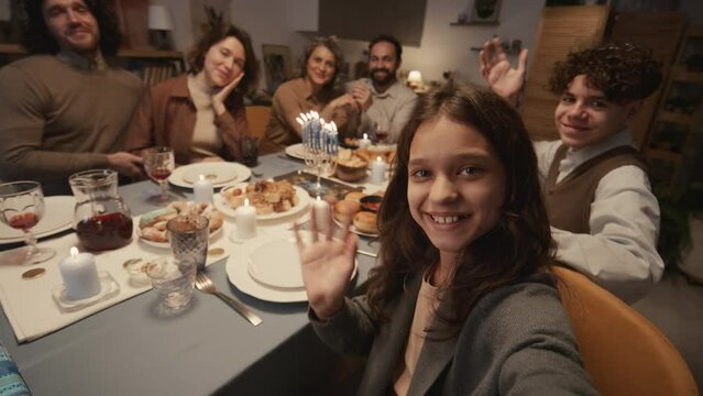 Medium shot of Jewish family of six sitting around dinner table with traditional foods, celebrating Hanukkah, teenage girl taking selfie shot on smartphone, everybody smiling and waving