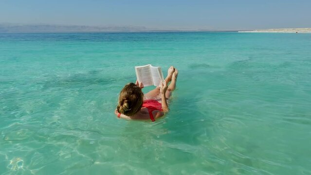 Beautiful Young Woman Floats in the Dead Sea in Red Swimsuit while Reading Book