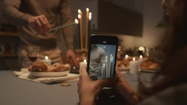 Medium shot of young Jewish man standing at table with festive foods, carefully lighting up candles on hanukkiah, and hands of young woman holding smartphone and filming him