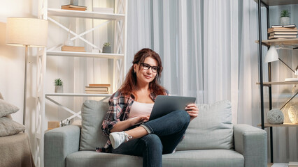 Virtual life. Happy woman. Home rest. Smiling middle-aged lady in spectacles sitting sofa scrolling laptop in light room interior.