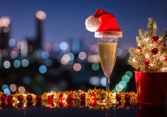 A glass of white wine that have santa claus hat on it with Christmas ornament decoration put on table with city bokeh light background. Christmas dining concept.