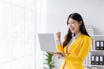 Excited cheerful asian business woman using laptop computer in the office, getting good news,...