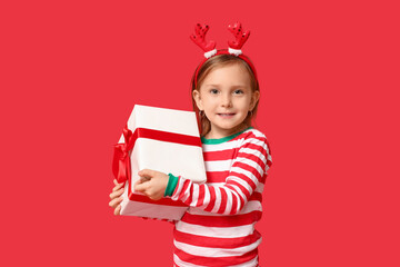 Cute little girl in pajamas with Christmas gift on red background