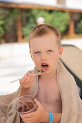 Child wrapped in three towels sitting on a sun lounger by the pool and eating ice-cream