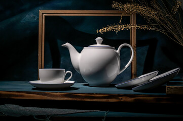 Cup and saucer, kettle, empty picture frame on background. Blue dark background. Conceptual still life.