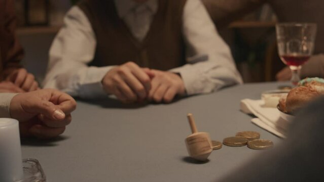 Close-up shot of hands of unrecognizable Jewish family sitting together, spinning dreidl and getting Hebrew letter Hei, meaning half, while celebrating Hanukkah at home