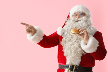 Santa Claus with tasty burger pointing at something on pink background