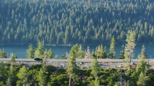 Driving by beautiful mountain road. Road trip concept. Mountain forest lake landscape Car driving through pine forest. Aerial view car driving through pine forest along lake shore in golden sunshine