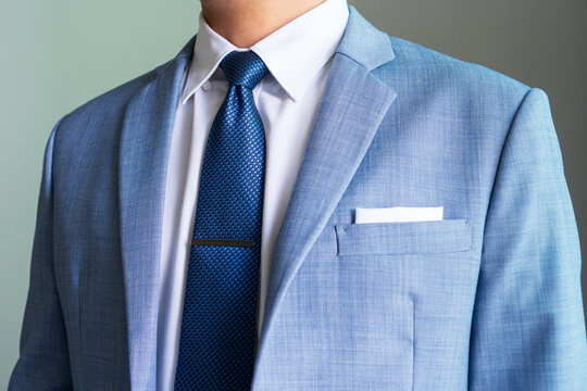 ฺBlue necktie in half winsor knot with single dimple and tie clip pairing with hidden button collar shirt and light blue blazer and white pocket square folded in presidential style.