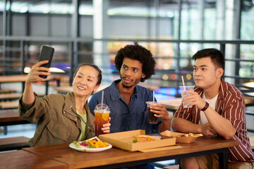 Smiling young woman taking selfie with friends when they are having soft drinks and pizza in cafe