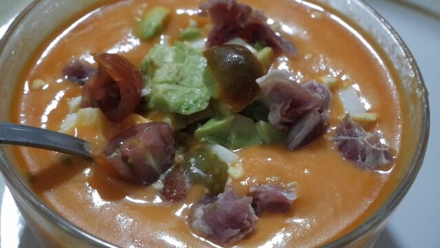 Bowl of gazpacho, traditional tomato soup from Spain. Toppings floating on top, cherry tomatoes, avocado, boiled egg, and ham slices.