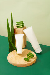 Front view of unbranded cosmetic tubes are displayed on a green table with a wooden platform and fresh aloe vera. Aloe vera effectively prevents and limits acne.