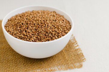 Organic dry buckwheat groats in a white bowl on a burlap napkin on a light background. Rural style. Vegetarian and vegan food. Concept of healthy eating. Horizontal orientation. Selective focus