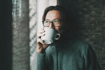One man with thoughtful and serene expression on face drinking coffee or tea alone at home looking...