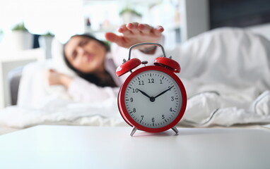Sleepy young woman portrait with one opened eye trying kill alarm clock. Early wake up not getting enough sleep going work concept. Female stretching hand to ringing alarm willing turn it off - obrazy, fototapety, plakaty