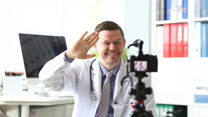 Smiling male millennial doctor wave his arm to camera saying hello to subscribers portrait