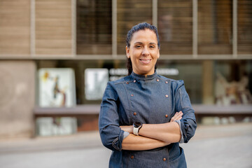 Female chef wears blue coat, broadly smiles and poses with arms crossed outdoors on the street. Restaurant worker, culinary gourmet, pastry chef.