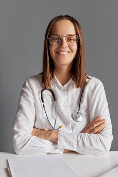 Pleased friendly female professional doctor in glasses and white medical lab coat looking at camera sitting at office desk against gray wall doing her paper work enjoying her job.