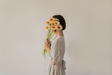 Beautiful woman in linen dress holding pastel gerber flowers bouquet over white wall. Aesthetic...