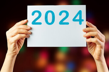 2024, Woman holding cardboard with number 2024 on background of faded lights. Happy New Year