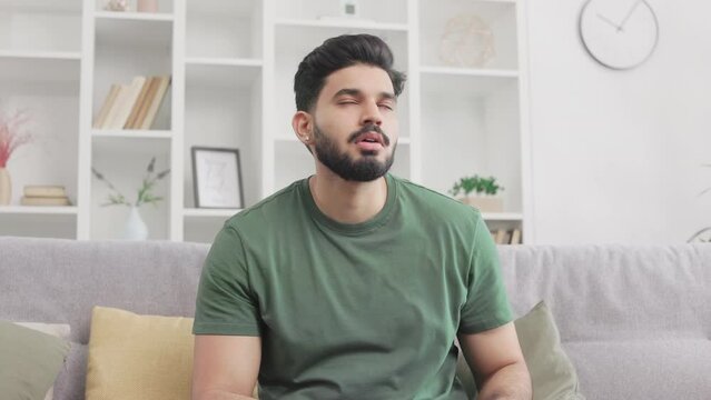 Desperate indian guy nervously tapping on smartphone screen while sitting on sofa at living room. Bearded guy in green t-shirt touching forehead and feeling frustration because damages on gadget.